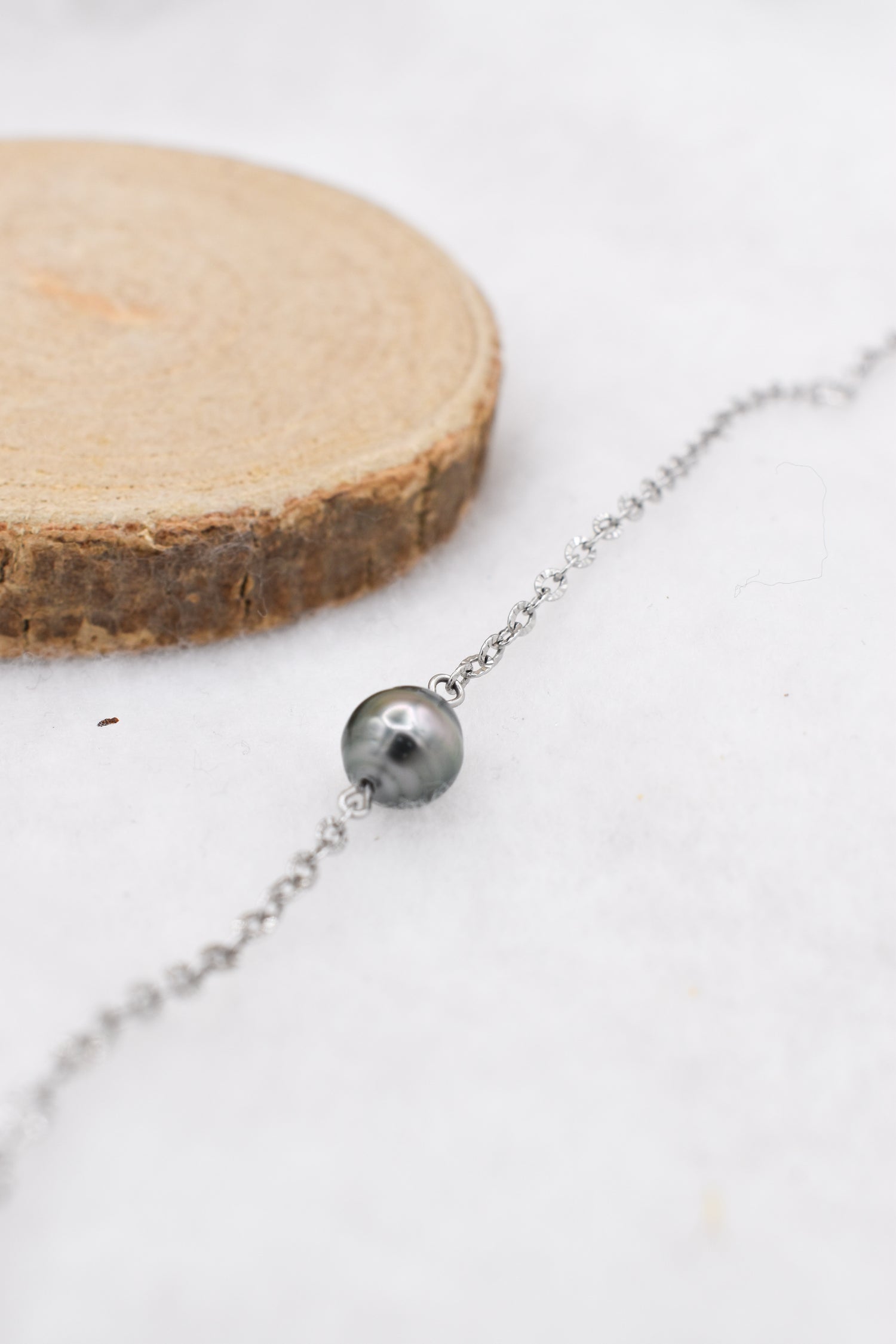 Bracelet minimaliste en chaine fine acier inoxydable et sa petite perle de Tahiti, posé sur un fond blanc et près d’une rondelle de bois naturelle.