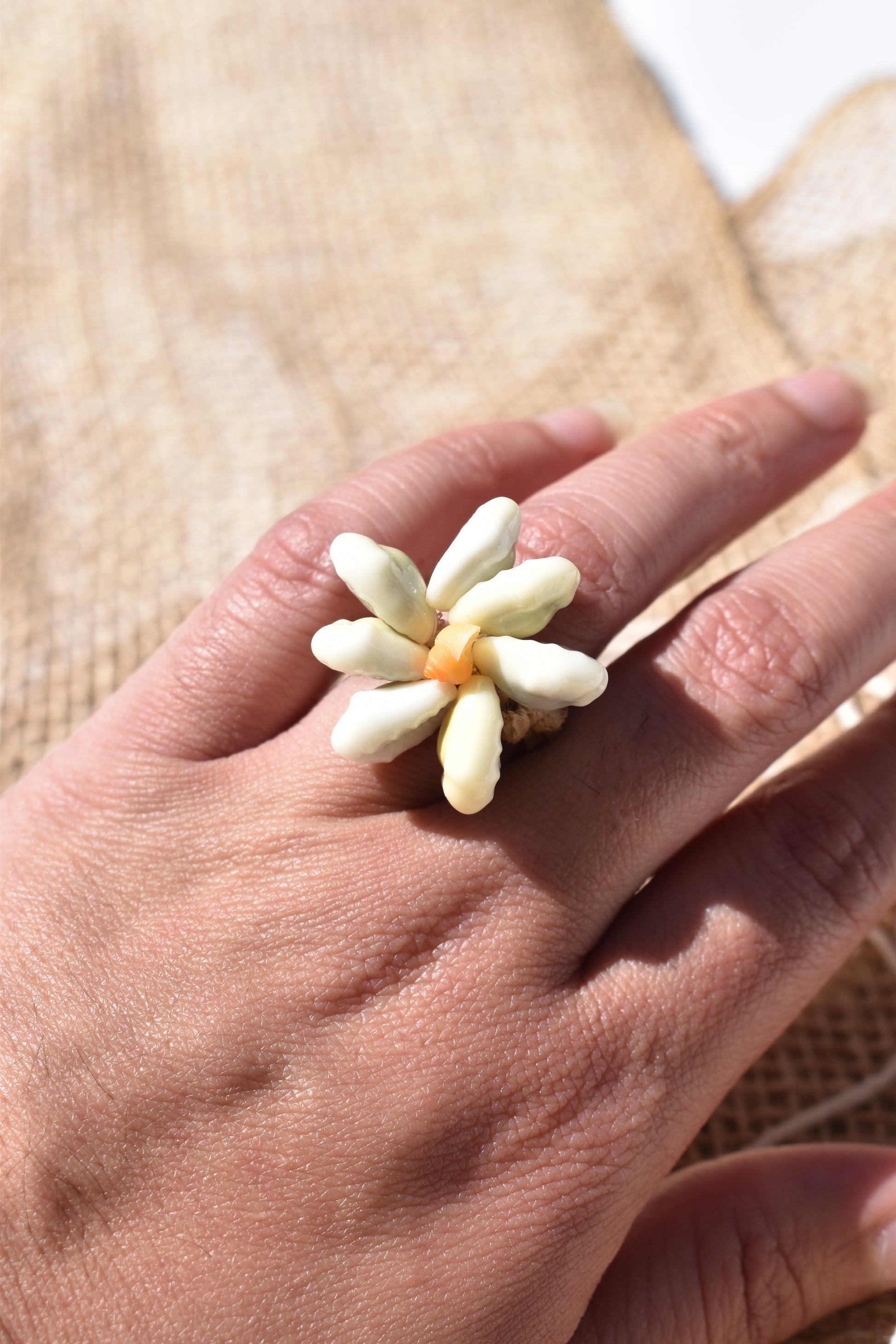 Bague en raphia tressé t coquillages naturel en forme de fleur, vu sur une main sur fond de nappe de fibre naturel , savoir faire de la Polynésie Française.